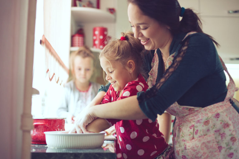 A woman and a young girl smile as they bake together in a kitchen, mixing ingredients in a bowl, while another child stands in the background. The scene looks warm and cheerful.
