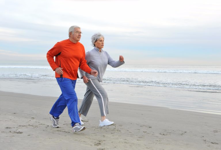 An older man and woman power walk along a sandy beach by the ocean, both wearing athletic clothing. The sky is overcast and the water is calm in the background.