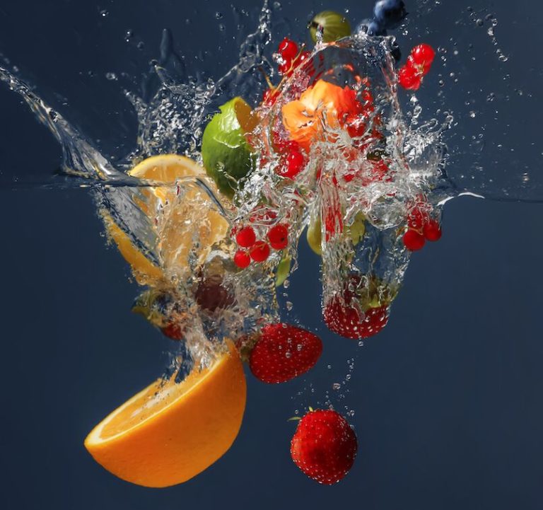 Various fruits, including an orange slice, strawberries, red currants, and a green apple, splash into clear water, creating dynamic splashes and bubbles against a dark background.