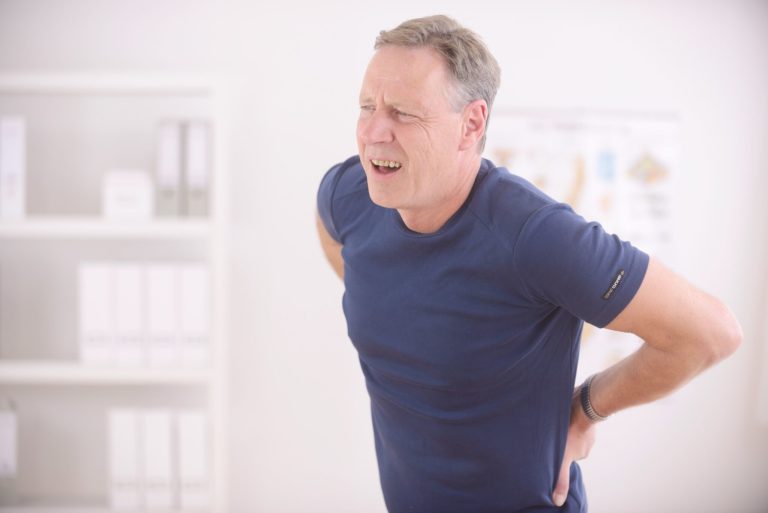 A middle-aged man in a blue shirt stands indoors, holding his lower back with both hands and grimacing in pain, suggesting he is experiencing back discomfort. Shelves with binders are visible in the background.
