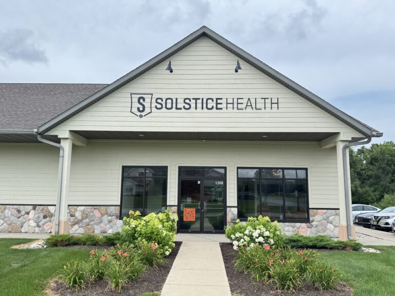 A beige building with a stone foundation and a sign reading Solstice Health above the entrance, surrounded by landscaped bushes and flowers, under a cloudy sky.