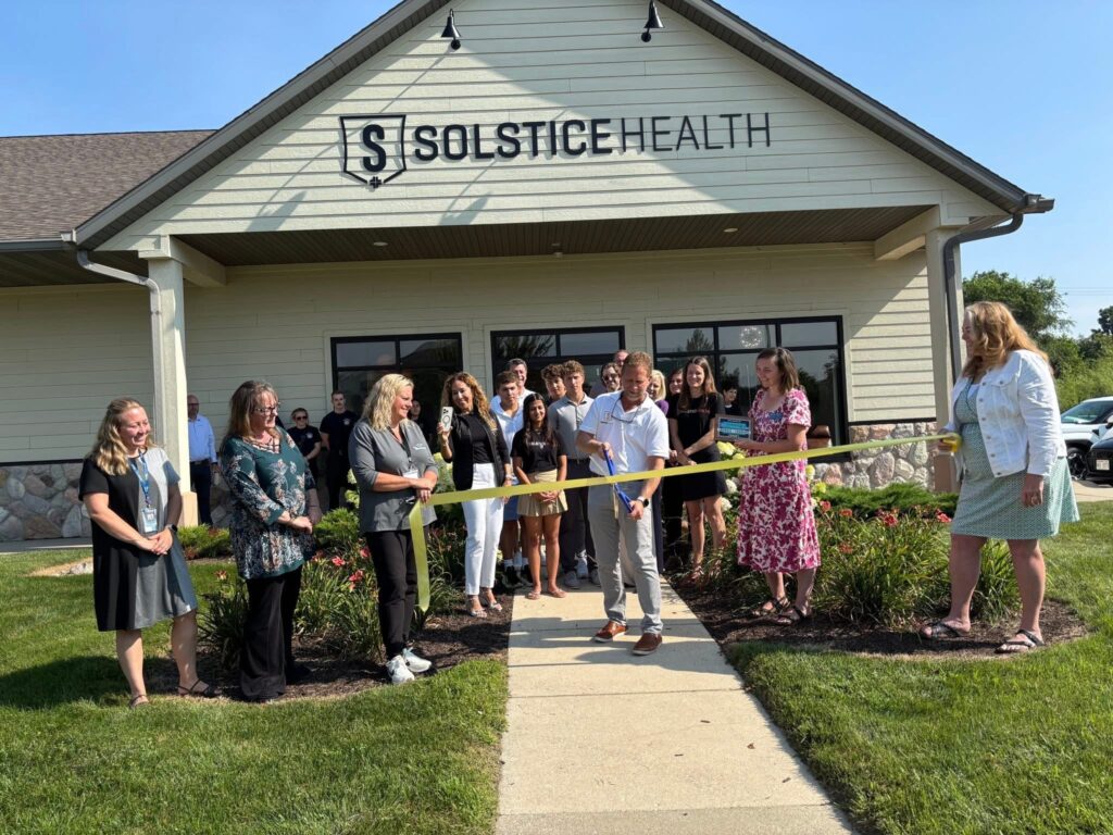 A group of people stand outside Solstice Health for a ribbon-cutting ceremony. A man in front cuts a yellow ribbon with large scissors, while others smile and watch, with the clinic building in the background.