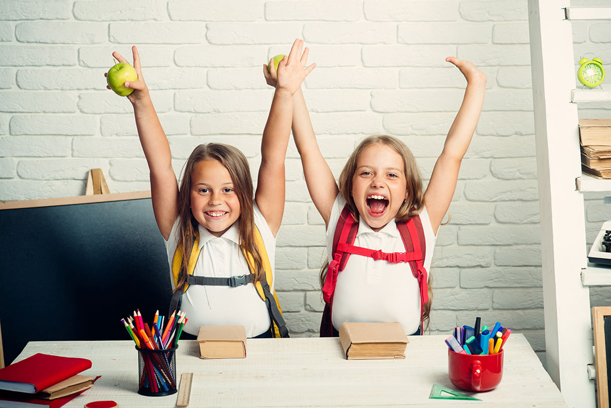 Two cheerful young girls with backpacks sit at a desk, each holding up a green apple and smiling with excitement. The desk has school supplies, and a chalkboard is in the background.