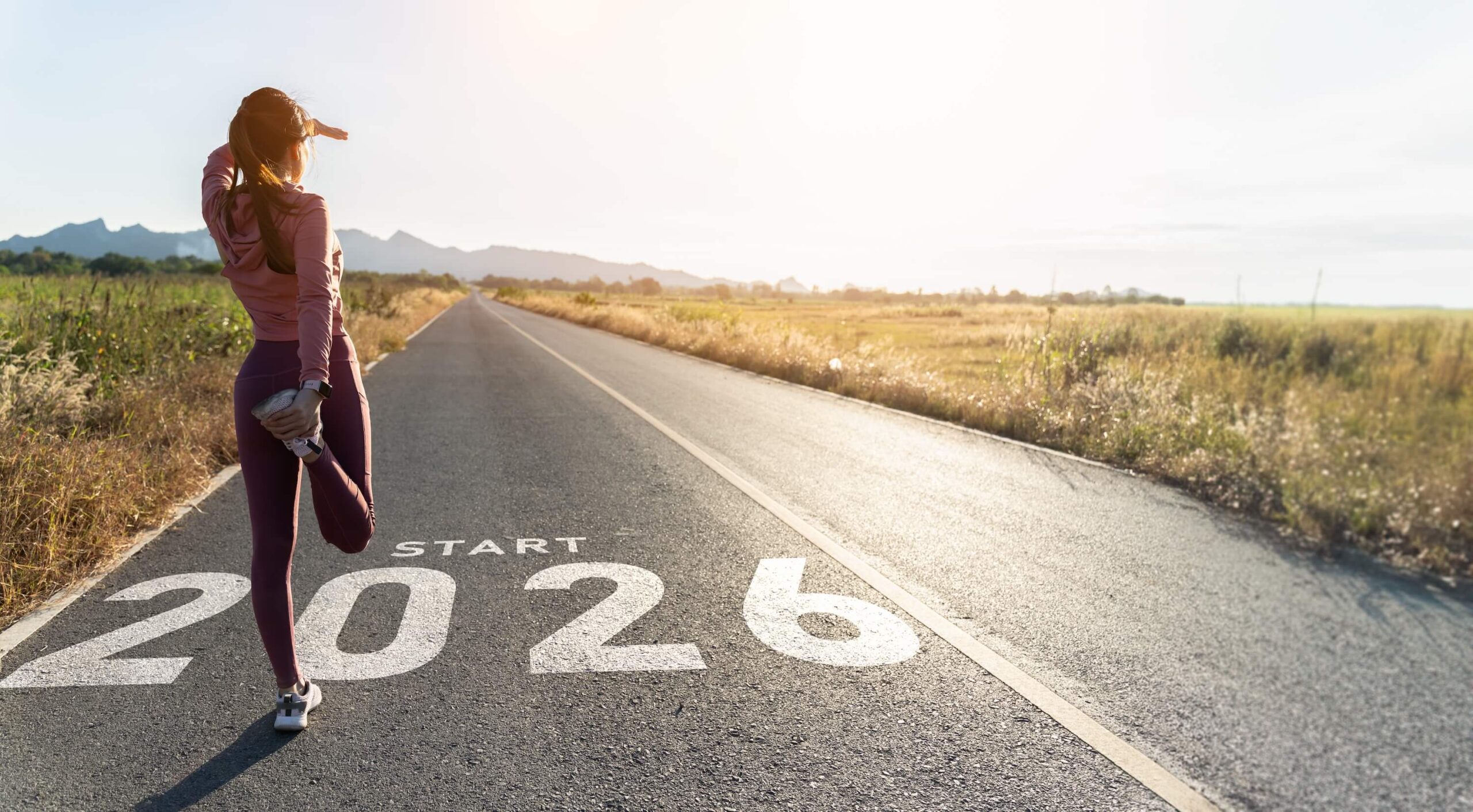 A woman in athletic clothing stretches on a rural road at sunrise, with START 2026 painted on the pavement, symbolizing a new beginning or goal ahead.