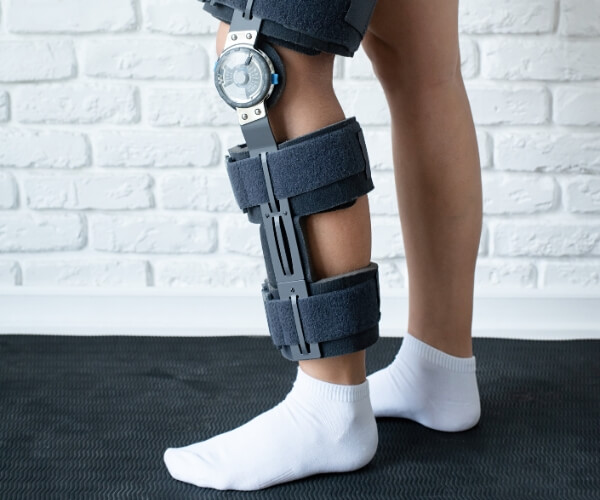 A person wearing a knee brace on their right leg stands on a black mat, wearing white socks. The adjustable knee brace, often recommended through Direct Primary Care, features sturdy straps and a metal hinge. The background is a white brick wall.