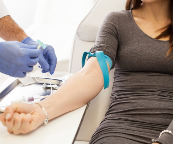 A person with rolled-up sleeves sits while a healthcare worker in gloves prepares to draw blood from their arm using a needle and tourniquet.